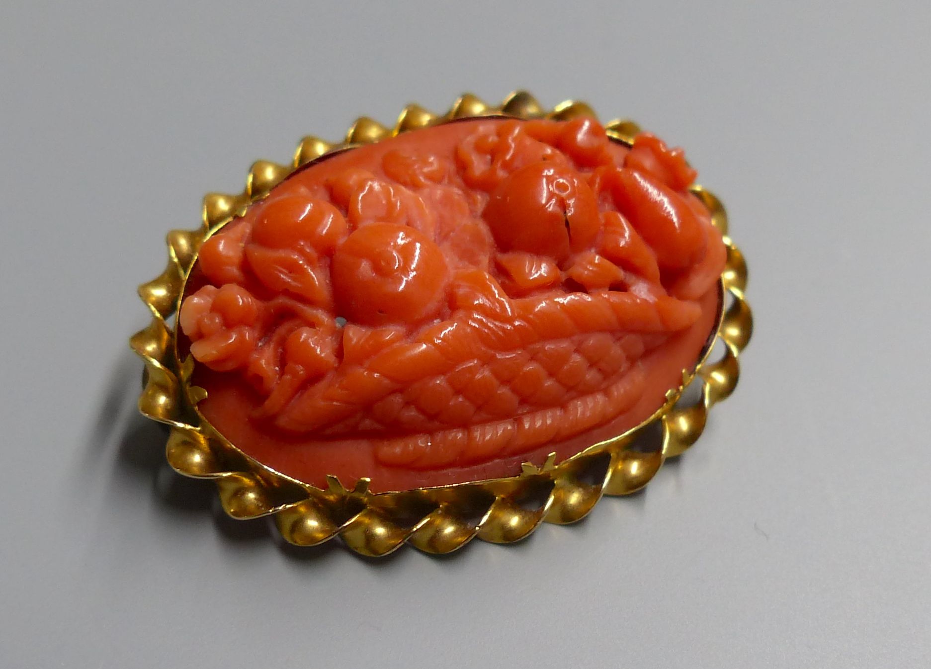 An antique yellow metal mounted oval coral set brooch, carved with a basket of flowers and fruit, 33mm, gross 6.4 grams.